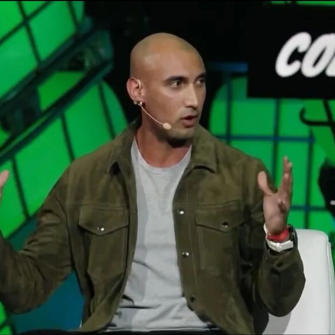 A bald man wearing a brown jacket and grey shirt speaks on stage with a headset microphone, gesturing with both hands in front of a green geometric background.
